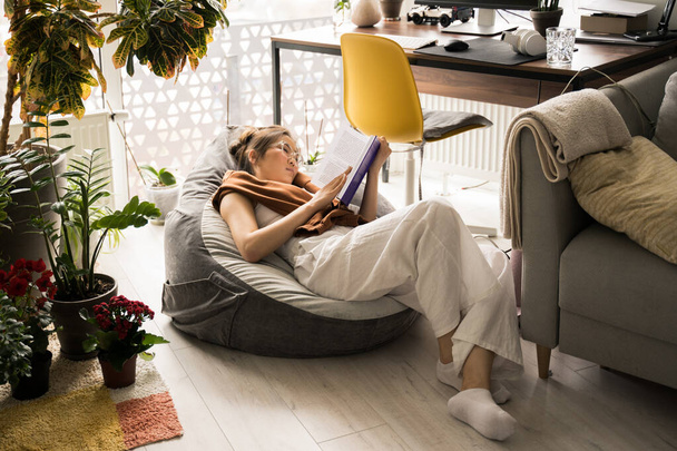 Woman wearing glasses relaxing on bag chair and reading book - Photo, image