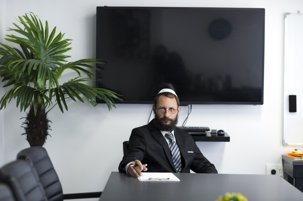 Ein jüdischer Mann mit Garderobe und Brille sitzt an einem Tisch im Büro. Jude in weißem Hemd, Businessanzug und Nationalhut Kippa und blickt ernst durch seine Brille, die Stirn gebeugt. - Foto, Bild