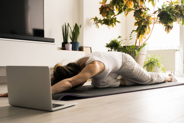 Woman stretching her back while training at the mat in front of the laptop - Foto, Bild