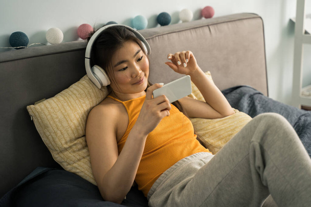 Woman wearing headphones laying at bed, listening music and looking at her smartphone - Foto, afbeelding