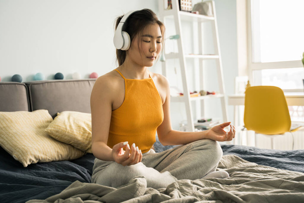 Woman wearing headphones sitting at home in the bed in lotus position, while meditating - Фото, изображение