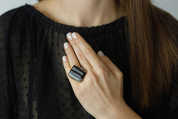 Beautiful stylish woman with modern square black ring on hand and white manicure, closeup. Fashionable female in black dress with unusual fused glass accessory. Beauty and care. - Photo, Image