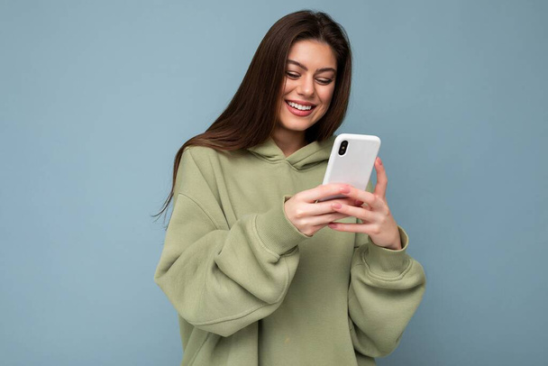 Lächelnd Schöne junge brünette Frau trägt stylischen grünen Kapuzenpulli mit Handy SMS schreiben isoliert auf dem Hintergrund Blick auf Smartphone-Bildschirm - Foto, Bild