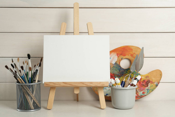 Easel with blank canvas, brushes, paints and palette on white wooden table - Fotografie, Obrázek