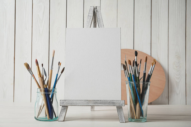Easel with blank canvas and brushes on white wooden table - Fotografie, Obrázek