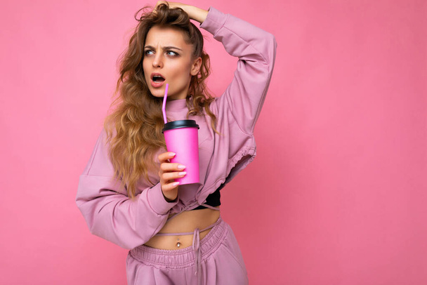 Foto de una hermosa joven rubia conmocionada con ropa deportiva rosa aislada sobre fondo rosa sosteniendo una taza de café de papel para beber y mirando hacia un lado. - Foto, imagen