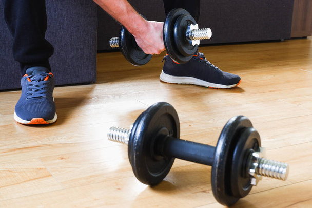 Home workout. Unrecognizable man in black leggings and sneakers doing strength training with dumbbells at home. - Foto, Imagen