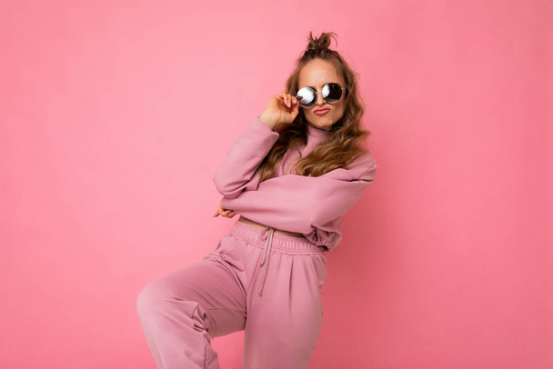 Photo of happy positive cool young beautiful blonde wavy-haired woman with sincere emotions wearing stylish pink sportswear and sunglasses isolated on pink background with copy space and having fun - Fotoğraf, Görsel