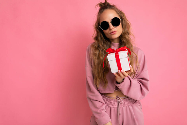 Shot of attractive serious calm young blond curly woman isolated over pink background wall wearing pink sport clothes and modern sunglasses holding gift box looking at camera - Photo, Image