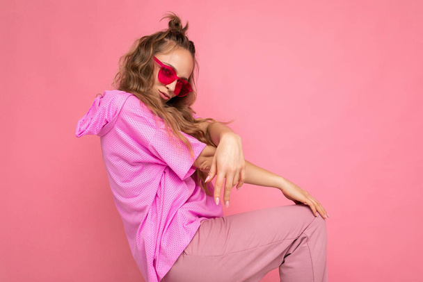 Charming positive young blonde curly woman isolated on pink background wall wearing casual pink t-shirt and stylish colourful sunglasses looking at camera and dancing - 写真・画像