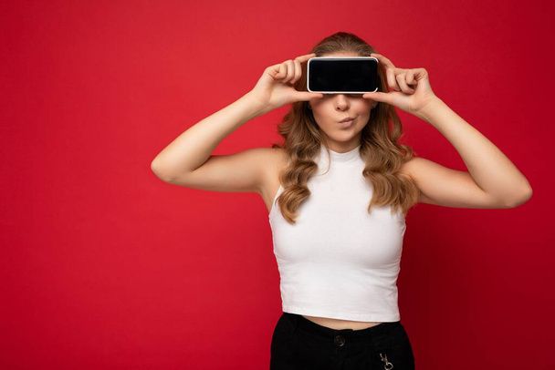 funny young blonde woman wearing white t-shirt isolated on red background with copy space holding smartphone showing phone in hand with empty screen for cutout - Fotó, kép
