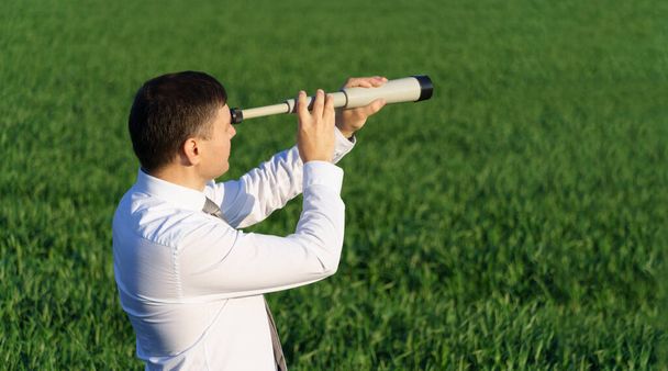 podnikatel pózuje dalekohledem, dívá se do dálky a hledá něco, zelenou trávu a modrou oblohu jako pozadí - Fotografie, Obrázek