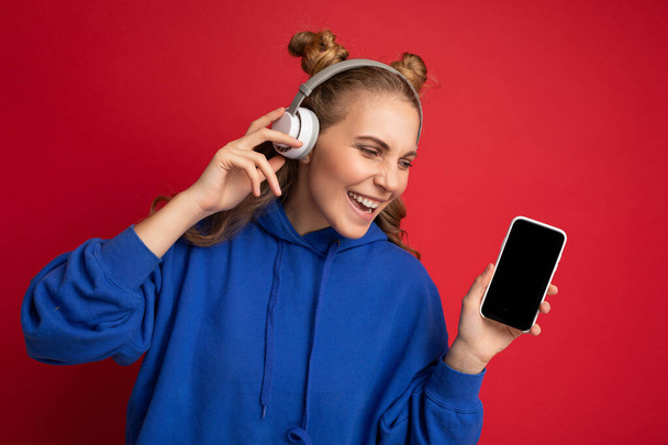 Vonzó pozitív mosolygós fiatal nő visel elegáns alkalmi ruhát elszigetelt színes háttér fali gazdaság és mutatja a mobiltelefon üres képernyőn kivágott visel fehér bluetooth - Fotó, kép