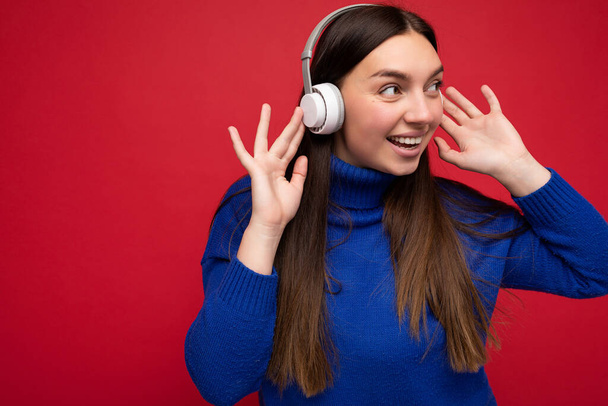 Photo of beautiful happy smiling young brunette woman wearing blue sweater isolated over red background wall wearing white bluetooth headphones listening to cool music and looking to the side - Photo, Image