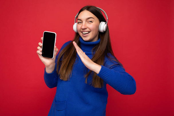 Vonzó pozitív mosolygós fiatal nő visel elegáns alkalmi ruhát elszigetelt színes háttér fali gazdaság és mutatja a mobiltelefon üres képernyőn kivágott visel fehér bluetooth - Fotó, kép