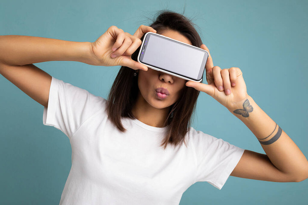 Photo of pretty positive young brunette woman good looking wearing white t-shirt standing isolated on blue background with copy space holding smartphone showing phone in hand with empty screen screen - Φωτογραφία, εικόνα