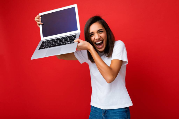 Foto van mooi Mooie vrolijke vrolijke jonge vrouw met kort donker brunet kapsel met computer laptop tot hoofd kijkend naar camera met wit t-shirt en jeans geïsoleerd over rode muur - Foto, afbeelding