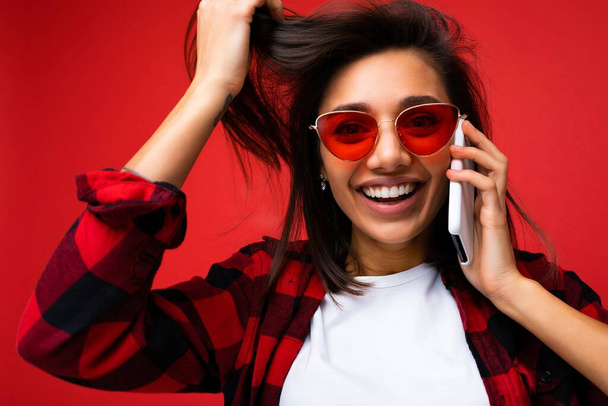Lähikuva kuva houkutteleva hymyilevä positiivinen nuori brunetti nainen yllään tyylikäs punainen paita valkoinen t-paita ja punainen aurinkolasit eristetty yli punainen tausta kommunikoida matkapuhelimeen katsoen kameraa - Valokuva, kuva