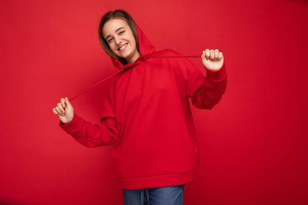 Photo shot of attractuve positive happy smiling brunette little girl wearing trendy red hoodie standing isolated over red background wall looking at camera - Photo, Image