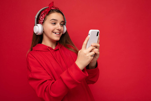 Κοντινό πλάνο πορτρέτο φωτογραφία του όμορφη ευτυχισμένη χαμογελαστή μελαχρινή κοπέλα φορώντας κόκκινη κουκούλα απομονώνονται σε κόκκινο φόντο εκμετάλλευση και τη χρήση smartphone επικοινωνία σε απευθείας σύνδεση φορώντας λευκά ασύρματα ακουστικά - Φωτογραφία, εικόνα