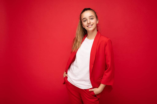 Φωτογραφία του όμορφου χαρούμενος χαμογελαστή μελαχρινή κοριτσάκι φορώντας μοντέρνο κόκκινο σακάκι και λευκό t-shirt για mockup στέκεται απομονωμένη πάνω από το κόκκινο φόντο τοίχο κοιτάζοντας την κάμερα - Φωτογραφία, εικόνα