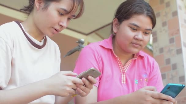 Kaksi thaimaalaista teini-ikäistä sisarta rento mekko nauttia chattailuun sosiaalisen viestinnän verkossa älypuhelimen istuessaan edessä talonsa. Aasialainen sisarus. Vaihdettavuus mobiiliteknologian kanssa. - Materiaali, video