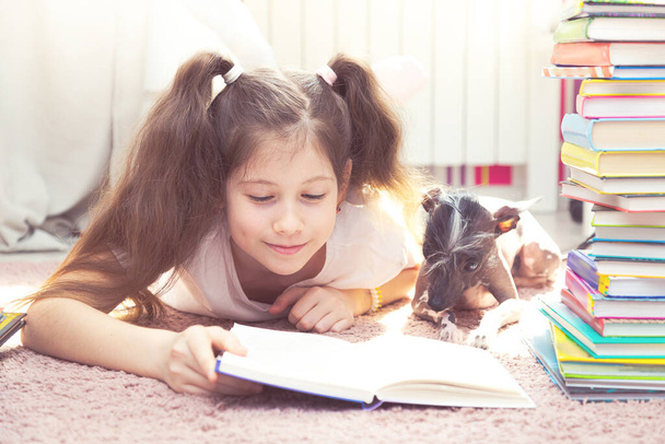 Little caucasian girl is playing with a dog on the floor. There are many books around them. The girl tries to read, the dog interferes with her. - Fotoğraf, Görsel