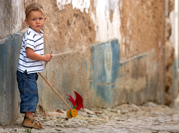 Cute 3-years old blond boy in casual clothes playing with handmade toy outdoors, old wall on background - Fotó, kép