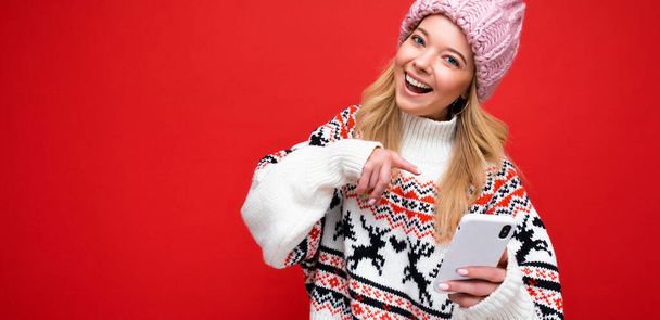 Panoramic Photo of beautiful smiling young blonde woman wearing warm knitted hat and winter warm sweater standing isolated over red background communicating online via smartphone looking at camera - Photo, image
