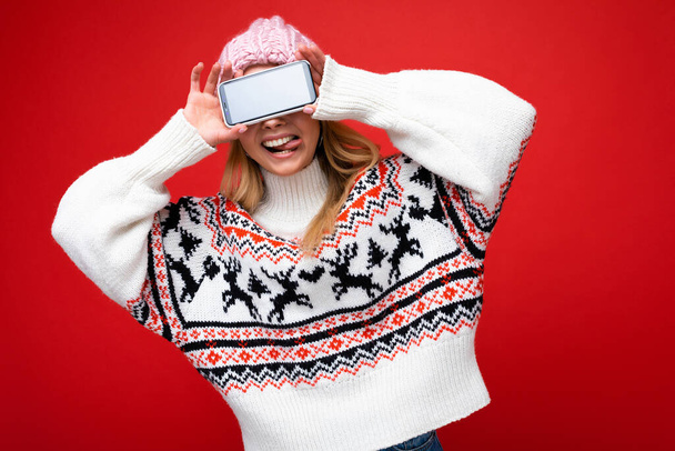 Foto de una atractiva joven rubia sonriente con un cálido sombrero de punto y un suéter cálido de invierno de pie aislado sobre un fondo rojo que muestra el teléfono inteligente con pantalla vacía para recortar divertirse y - Foto, imagen