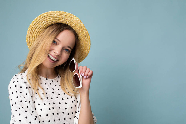 Foto záběr krásné pozitivní mladé blondýny žena na sobě letní ležérní oblečení a držení stylové sluneční brýle izolované přes barevné pozadí při pohledu na kameru - Fotografie, Obrázek