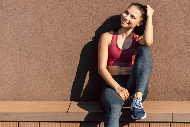 Image of an athlete female smiling, looking at the camera, resting after exercising outdoors on a sunny day. Sporty woman relaxing after aerobics in the morning outside. - Foto, Bild