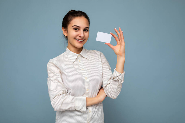 Shot van aantrekkelijke positieve glimlach jonge brunette vrouw het dragen van witte blouse geïsoleerd over blauwe achtergrond met credit card kijken naar camera - Foto, afbeelding