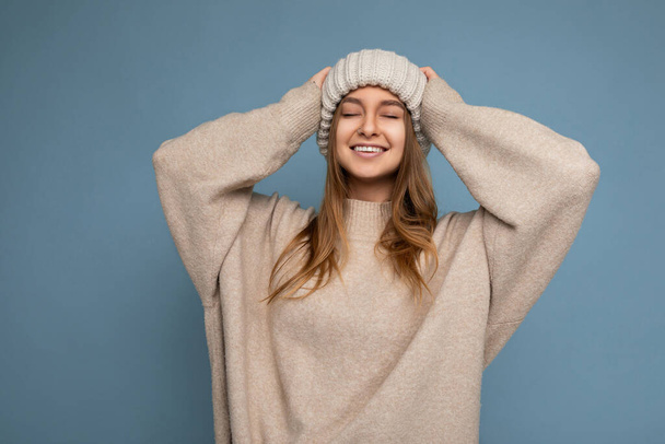 Съемка увлекательный счастливый улыбающийся взрослый темно-блондинка женщина изолированы на синем фоне стены в бежевом теплый свитер и вязаные бежевые шляпы наслаждаясь с закрытыми глазами - Фото, изображение