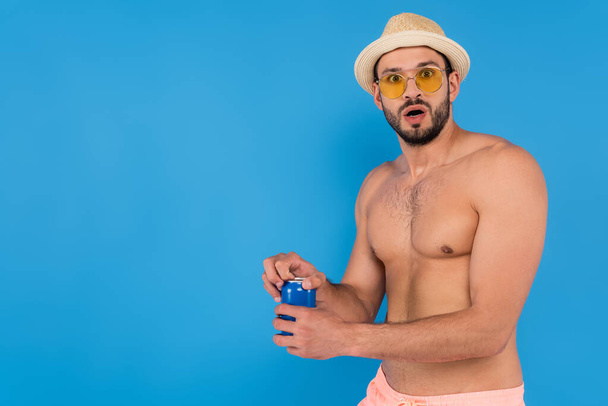 Emocionado hombre sin camisa con sombrero de paja y gafas de sol sosteniendo bebida enlatada aislada en azul  - Foto, Imagen