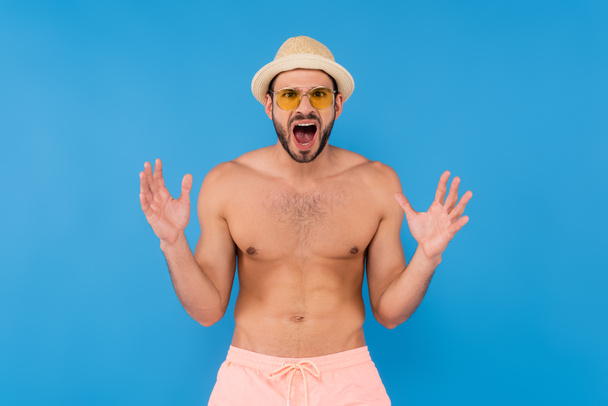 Wütender Mann mit Sonnenbrille und Sonnenhut schreit vereinzelt auf blauem Grund  - Foto, Bild