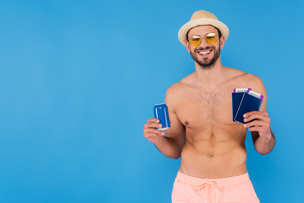 Hombre alegre sin camisa en gafas de sol sosteniendo bebida enlatada y pasaportes aislados en azul  - Foto, imagen