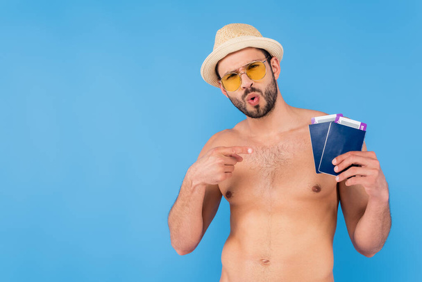 Homme excité avec des lunettes de soleil pointant vers les passeports avec des billets d'avion isolés sur bleu  - Photo, image
