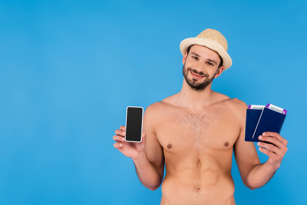 Shirtless man in sun hat holding smartphone and passports isolated on blue  - Foto, afbeelding