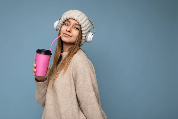 Beautiful sexy adult blond woman wearing knitted hat beige sweater and white wireless earphones listening to music isolated over blue background holding paper coffee cup for mockup drinking and - Foto, afbeelding