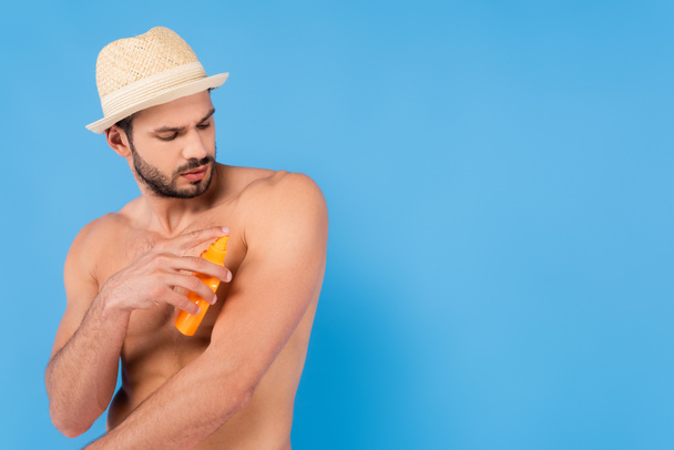 Muscular man in sun hat applying sunscreen isolated on blue  - Photo, Image