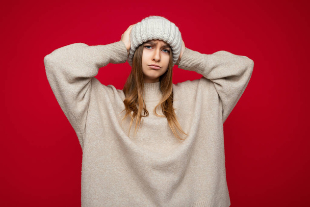 Foto de bela triste chateado insatisfeito jovem loira escura de pé isolado sobre a parede de fundo vermelho vestindo suéter bege quente e chapéu bege de inverno olhando para a câmera - Foto, Imagem