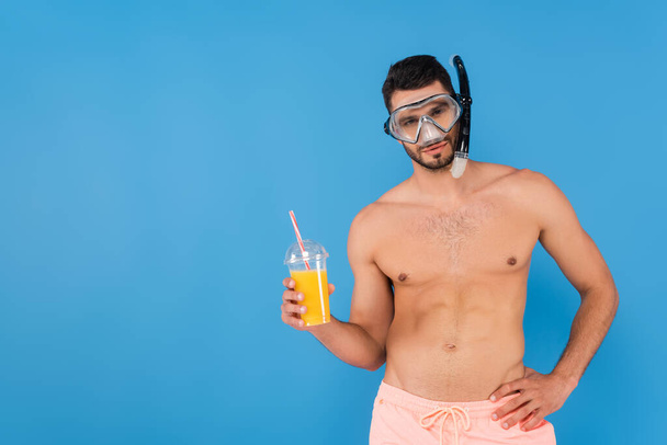 Shirtless man in swimming goggles holding orange juice isolated on blue  - Foto, afbeelding
