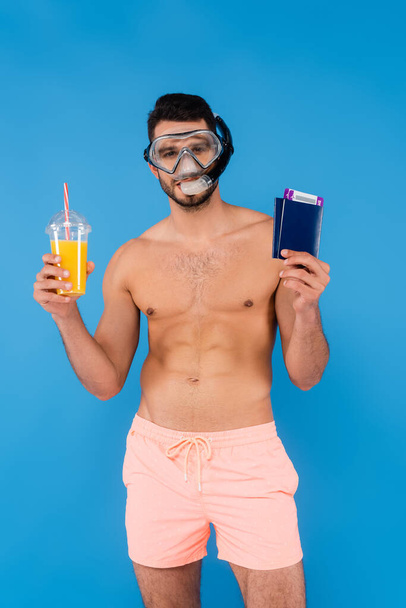 青い上に隔離されたパスポートとオレンジジュースを保持している水泳ゴーグルの白人男性  - 写真・画像