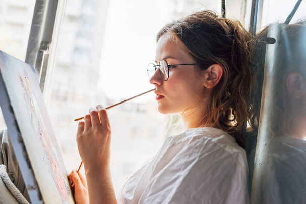 Oldalnézet egy csinos női művész festmény vászonra a művészeti stúdióban ül az ablak mellett. Egy női festő szemüveges festmény olajjal keresi a képzelet a műhelyben. - Fotó, kép