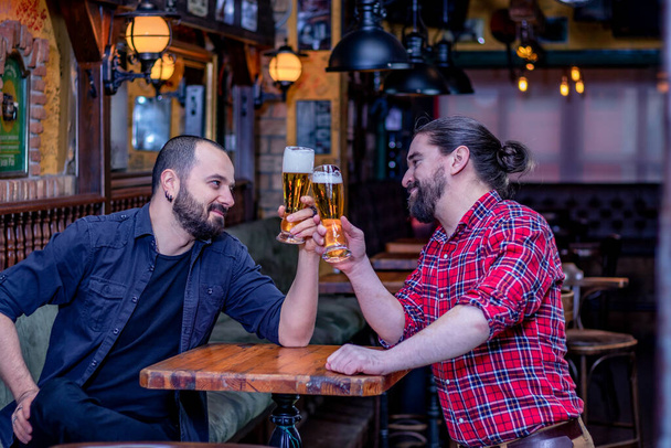 Δύο άντρες φίλοι πίνουν στο καφέ. Γελάνε και κάνουν πρόποση με ψηλά ποτήρια γεμάτα μπύρα. Πίσω στην παμπ. Εις υγείαν. - Φωτογραφία, εικόνα