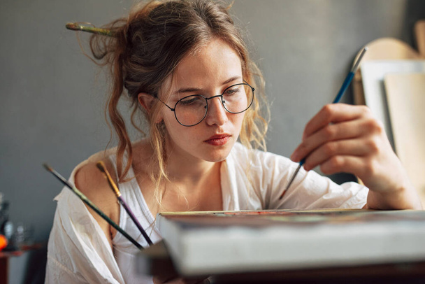Portrait of an artist female standing next to the easel with canvas painting something in her art studio. A professional young woman painter in transparent spectacles draws in the workshop. - Photo, image