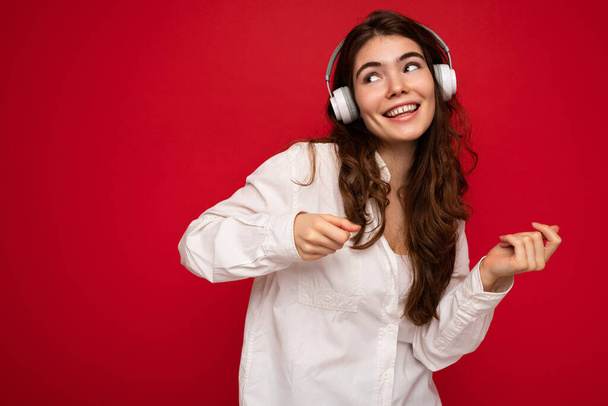 Aantrekkelijke vrolijke glimlach jonge brunette vrouwelijke persoon met een wit shirt en optische bril geïsoleerd over rode achtergrond dragen witte draadloze bluetooth oortelefoons luisteren naar muziek en dansen - Foto, afbeelding
