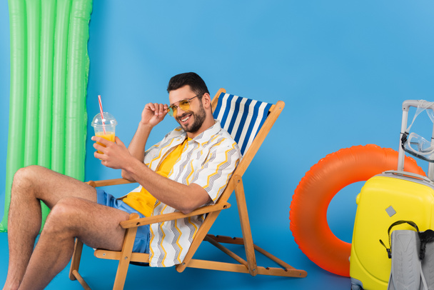 Hombre sonriente con jugo de naranja sentado en la silla de cubierta cerca de la maleta, aletas de natación y anillo inflable sobre fondo azul  - Foto, imagen