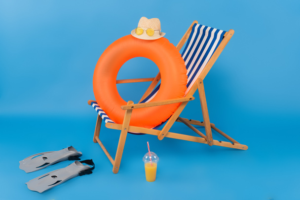 Sombrero de paja en anillo inflable cerca de aletas de natación y jugo de naranja sobre fondo azul  - Foto, Imagen